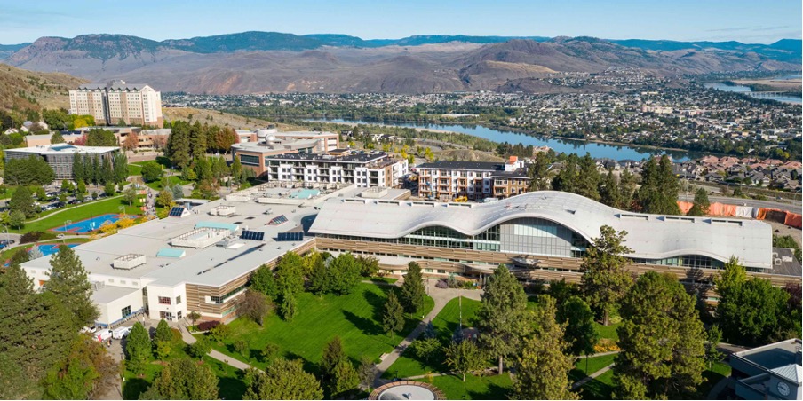 Aerial view of the Thompson Rivers University (TRU) campus in Kamloops, British Columbia. The image features modern academic and residential buildings surrounded by green spaces and trees. In the background, the Thompson River winds through the city of Kamloops, with rolling hills and distant mountains providing a scenic backdrop under a clear blue sky.








